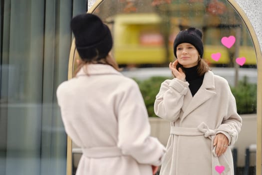 A woman stands in front of a large mirror, wearing a cozy coat and beanie. She smiles while looking at her reflection, surrounded by light and vibrant colors, creating a cheerful urban atmosphere.