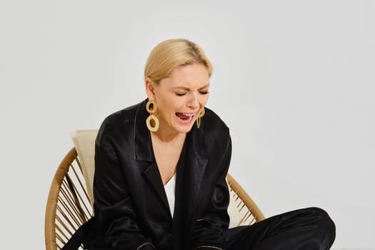 Upset woman sobs loudly while sitting on a wicker chair. Grief and despair are expressed on the woman's face. The minimalist background emphasizes her expression.