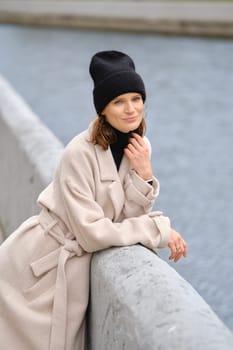 Cute woman leans casually against a railing beside a serene waterway, dressed in a stylish black beanie and a soft beige coat, enjoying the tranquil atmosphere of the urban landscape.