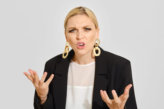 A woman stands indoors, visibly frustrated with raised hands and an expressive facial gesture. She wears a black blazer and large earrings, emphasizing her emotional state and strong presence.