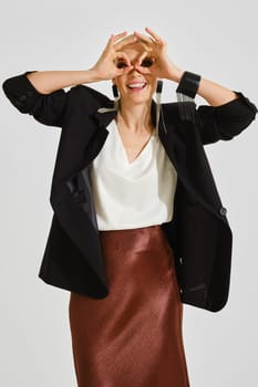 A woman is joyfully posing indoors, wearing a chic black blazer over a white top and a shimmering bronze skirt. She playfully forms glasses with her hands, showcasing her lively personality.