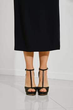 Unrecognizable woman stands in a sleek black dress, highlighting her elegant black high-heeled shoes in a bright, contemporary room. The polished floor reflects the chic footwear choice.