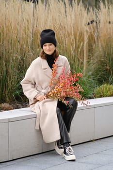In an urban park, a woman dressed in a light coat and beanie sits on a concrete bench, holding colorful autumn branches. The fall foliage and tall grasses create a serene setting around her.