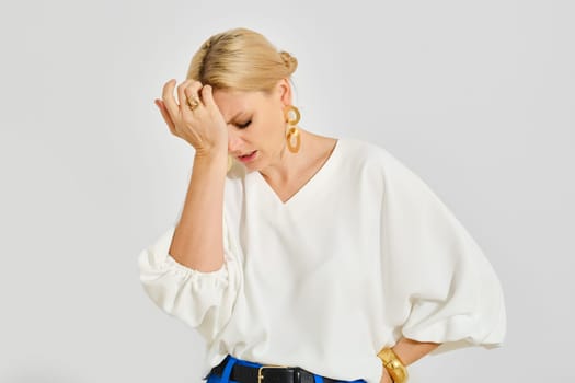 A woman with blonde hair wearing a white blouse stands with her hand on her forehead, visibly frustrated. Her expression conveys a sense of concern or disappointment in a neutral setting.