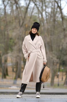 A woman dressed in a beige coat and black beanie poses confidently in a serene park setting. The trees show signs of autumn, creating a warm, inviting atmosphere while she carries a backpack.