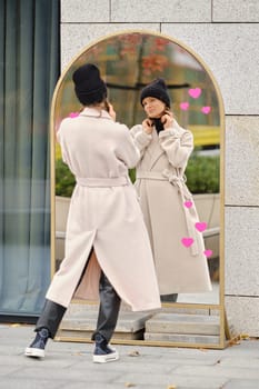 A woman in a fashionable coat and beanie adjusts her earrings while looking into a large mirror. The park\'s autumn foliage creates a cozy atmosphere, enhancing her reflective moment.