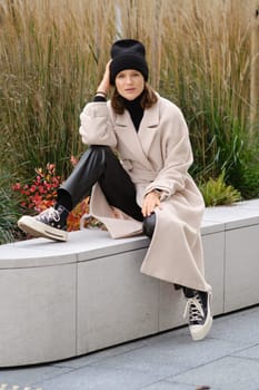 A young woman sits casually on a ledge, wearing a long beige coat and black beanie, surrounded by lush greenery and vibrant plants. She completes her look with black pants and stylish sneakers.