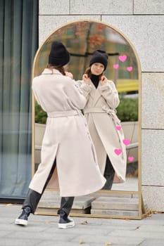 A woman stands outdoors, looking at her reflection in a modern mirror while wearing a coat and a beanie.