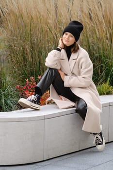 A woman sits comfortably on a stone bench, wearing a stylish coat and a beanie, with her chin resting on her hand. Surrounded by lush foliage and colorful plants, she exudes a relaxed vibe.