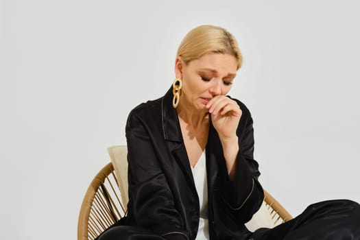 A crying woman in elegant black clothing sits thoughtfully in a stylish rattan chair, her expression revealing deep emotion.