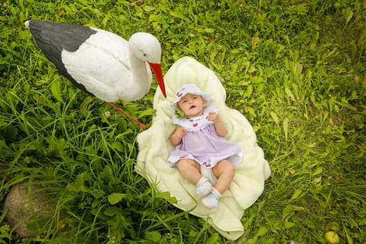 A cute little baby comfortably lies on a soft blanket, curiously gazing up at a stork perched beside her