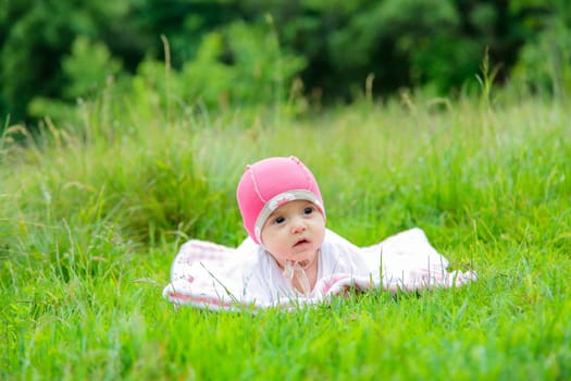 A sweet baby is blissfully lying on the green grass in a pink hat and really enjoying the wonderful view in the fresh air