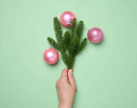 Female hand holding shiny pink balls and green spruce branches on green background, Christmas background