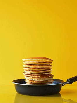 A tall stack of golden-brown pancakes sits inside a modern black frying pan, with a yellow background enhancing the warm colors of the breakfast scene