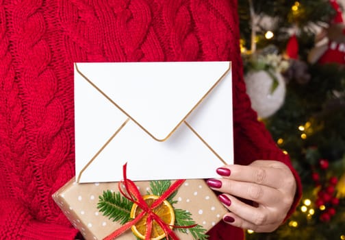 Woman in red sweater holding in hands with blank envelope against Christmas tree with lights, mockup, copy space, front view. Girl with red nails opening holiday invitation. Cozy holidays atmosphere