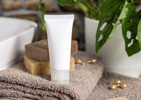 White cream squeeze tube on brown folded towels near vessel sink, soap bars and green plant in bathroom, close up, packaging mockup. Natural cosmetic product for skin care