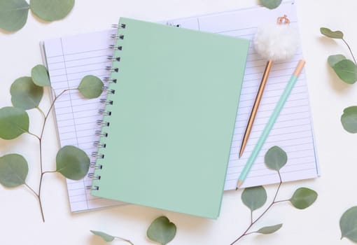 Light green spiral notebook on a white table near green eucalyptus leaves and pen top view, Blank school textbook mockup