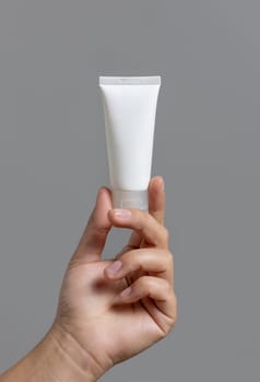 Female hand holding white squeeze cream tube against grey close up, cosmetic mockup, copy space. Skincare beauty product package, minimal composition