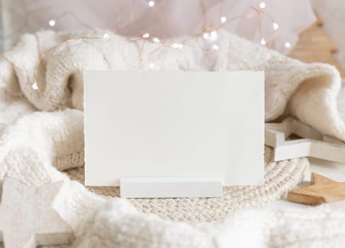 Horizontal blank paper card near white knitted sweater, wooden Christmas decor and lights, copy space. Winter atmospheric mock up, family greeting in rustic or scandinavian style