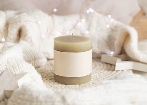 Pillar candle with blank label near white knitted sweater, wooden Christmas decor and lights, copy space. Winter atmospheric mock up in rustic or scandinavian style