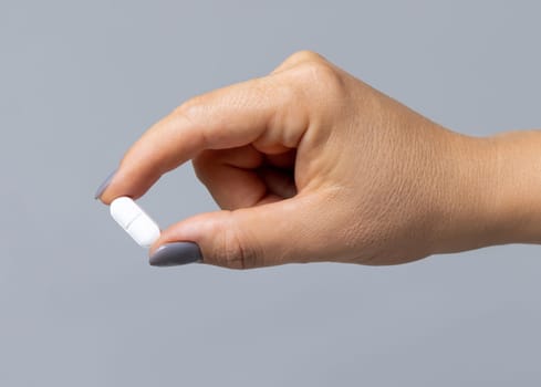 Female hand holding a white pill with fingers closeup against grey background. Healthcare, medical and pharmaceutical concept.