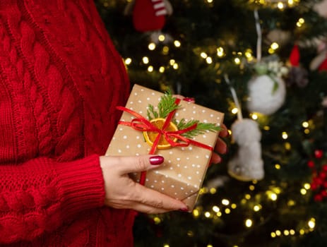Woman in red sweater with present in hands against Christmas tree lights, side view. Girl with red nails keeping wrapped gift box. Cozy holidays atmosphere