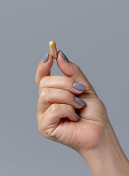 Female hand holding a yellow capsule with fingers closeup against grey background. Healthcare, medical and pharmaceutical concept, herbal alternative cure