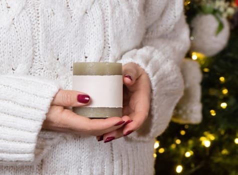 Woman in white sweater with cream pillar candle in hands against Christmas tree lights, mock up, copy spac. Candle with horizontal label in female hands. Holidays atmosphere