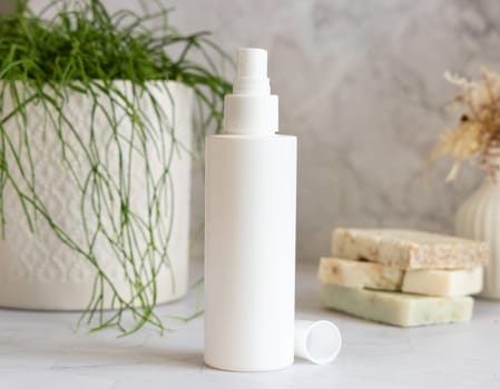 White spray cosmetic bottle on white marble against potted green plant and soap bars in bathroom, close up, packaging mockup. Natural product for facecare, toner or micellar water