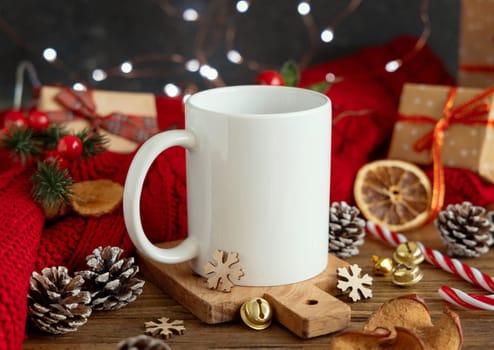 White blank coffee mug near red knitted sweater and Christmas decor against lights and wrapped presents, copy space. Warm winter family atmospheric mock up, Holiday time