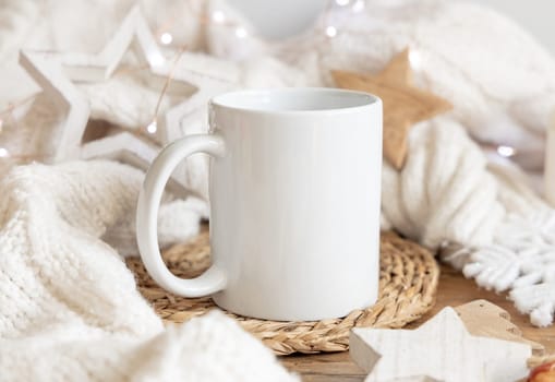 White blank coffee mug near white knitted sweater, wooden Christmas decor and lights, copy space. Winter atmospheric mock up in rustic or scandinavian style