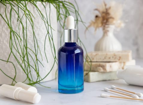 Blue glass dropper bottle on white marble against potted green plant and soap bars in bathroom, close up, packaging mockup. Cosmetic product for facecare, beauty routine