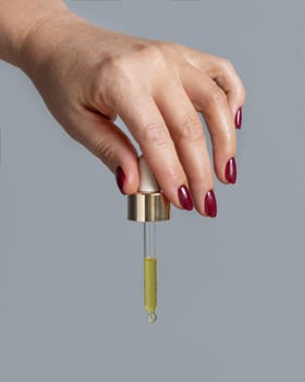 Female Hand with dark red nails holding with pipette filled with yellow liquid over grey close up. Natural skincare product, serum or essential oil. Medical research