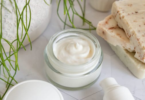 Opened cream jar with white blank lid on white marble against potted green plant and soap bars in bathroom, close up, packaging mockup. Cosmetic product for facecare, beauty routine