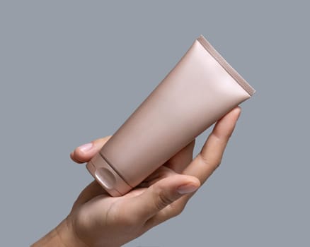 Female hand holding beige squeeze cream tube against grey close up, cosmetic mockup, copy space. Skincare beauty product package, minimal composition