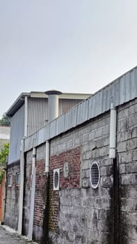 An industrial building partially hidden by a brick wall under a blue sky. The image captures urban architecture with a mix of materials and textures in a relaxed setting.