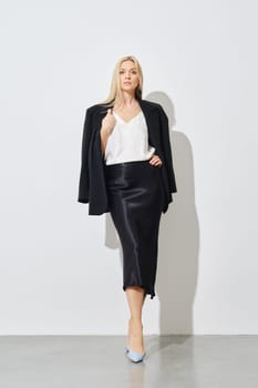 Stylish woman posing against white wall in a white blouse, a silk long skirt and a jacket draped over her shoulders