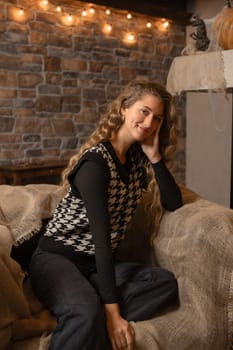 A young woman with long, wavy hair smiles softly while seated on a cozy couch draped with a burlap. The warm autumn light enhances the rustic charm of the stone wall behind her.