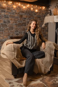 A woman sits comfortably on a couch draped with burlap in a cozy room decorated for autumn. Soft lighting casts a warm glow, creating a relaxed atmosphere perfect for unwinding.