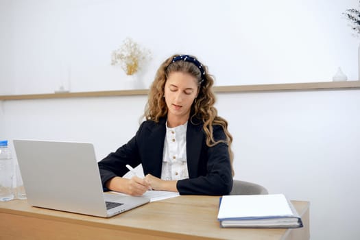 A young woman sits attentively at her modern office desk, writing notes and reviewing a laptop screen. Natural light fills the space as she engages in her work with determination and concentration.