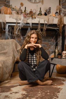 A woman sits comfortably on the floor with her hands under her chin, surrounded by Halloween decorations. The cozy room features pumpkins, cobwebs, and a welcoming autumnal atmosphere.