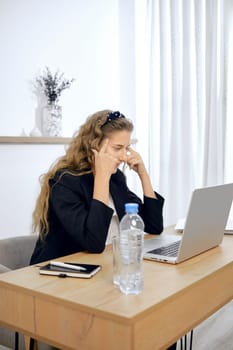 A young woman sits at a wooden desk in a well-lit room, focusing intently on her laptop screen. She appears deep in thought, with a water bottle and notebook nearby, likely engaged in work or study.