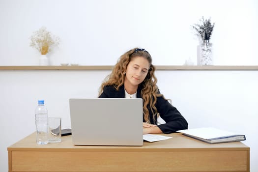 A young woman sits at a contemporary wooden desk, engaged in work on her laptop. Papers and a water bottle are placed nearby, creating a tidy workspace in a bright, minimalist office environment.
