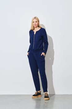 Blonde woman posing in knitted blue pantsuit against white wall