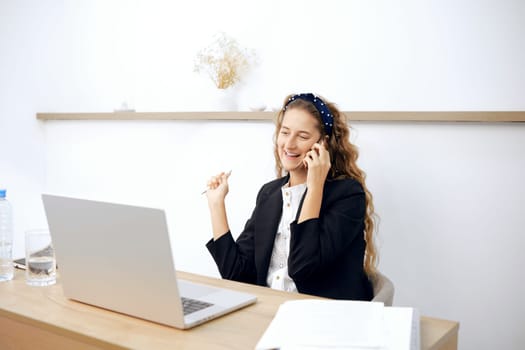 A young woman with long, curly hair smiles during a phone call and takes notes at her tidy desk. The bright office features light wood accents and minimal decor, creating an inviting workspace.