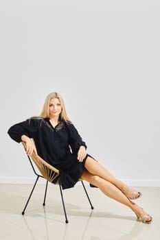 Fashionable woman in loose black dress and silver sandals posing on a modern chair
