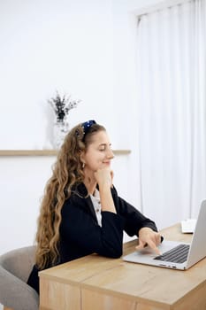 A woman with wavy hair sits at a wooden desk, focused on her laptop. She wears a black blazer and appears engaged in her work. The room is well-lit with white curtains and simple decor.