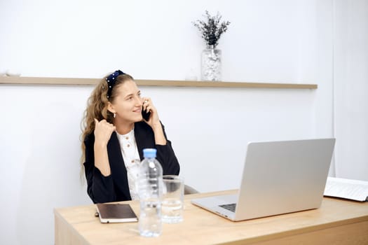 A woman with curly hair enjoys a lively phone conversation at her desk, surrounded by a laptop, water bottle, and notepad. The bright office space features minimalistic decor and a wooden shelf.