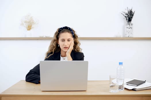 A young woman sits at a wooden desk, focused on her laptop while resting her chin on her hand. A water bottle and glass sit nearby, alongside notebooks and a notepad, in a bright, airy office.