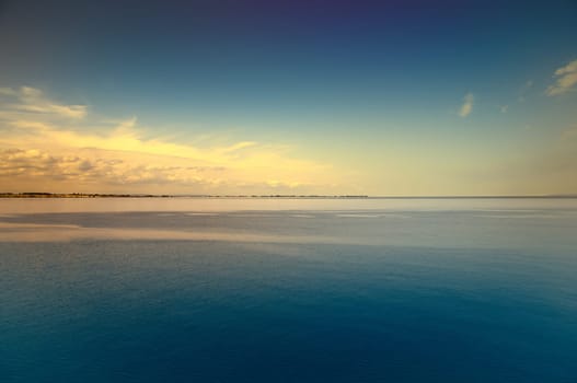 Tranquil seascape under a clear blue sky. The water reflects gentle clouds, while the horizon emphasizes the distance.